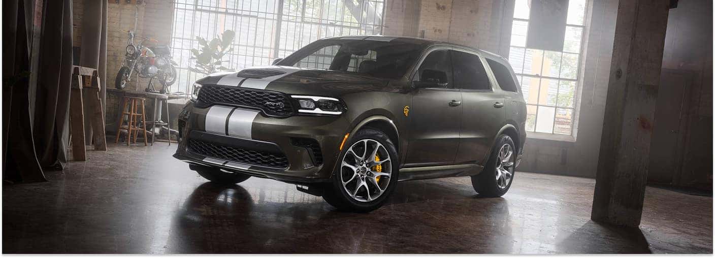 Dark green 2026 Dodge Durango with white racing stripes in a warehouse, front three-quarter view.