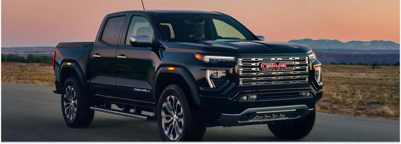 Black 2026 GMC Canyon Denali pickup truck driving on a road at sunset.