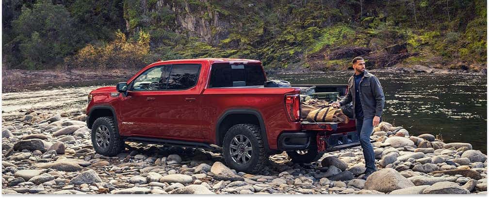 Red 2026 GMC Sierra 1500 AT4X pickup truck parked on a rocky riverbank with open tailgate.