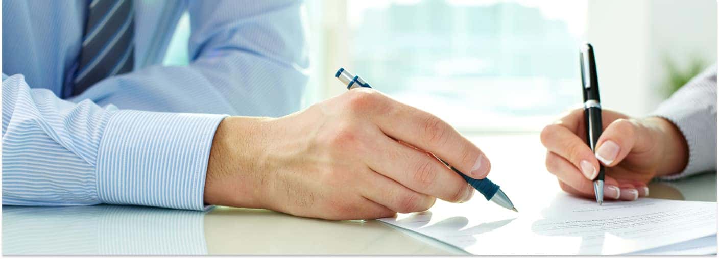 Two people signing auto financing paperwork at a desk with pens.
