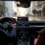 A couple is driving a 2025 Mazda CX-5 with hands on the wheel and showing instrument panel and gauges along with interior features while looking out of windshield.