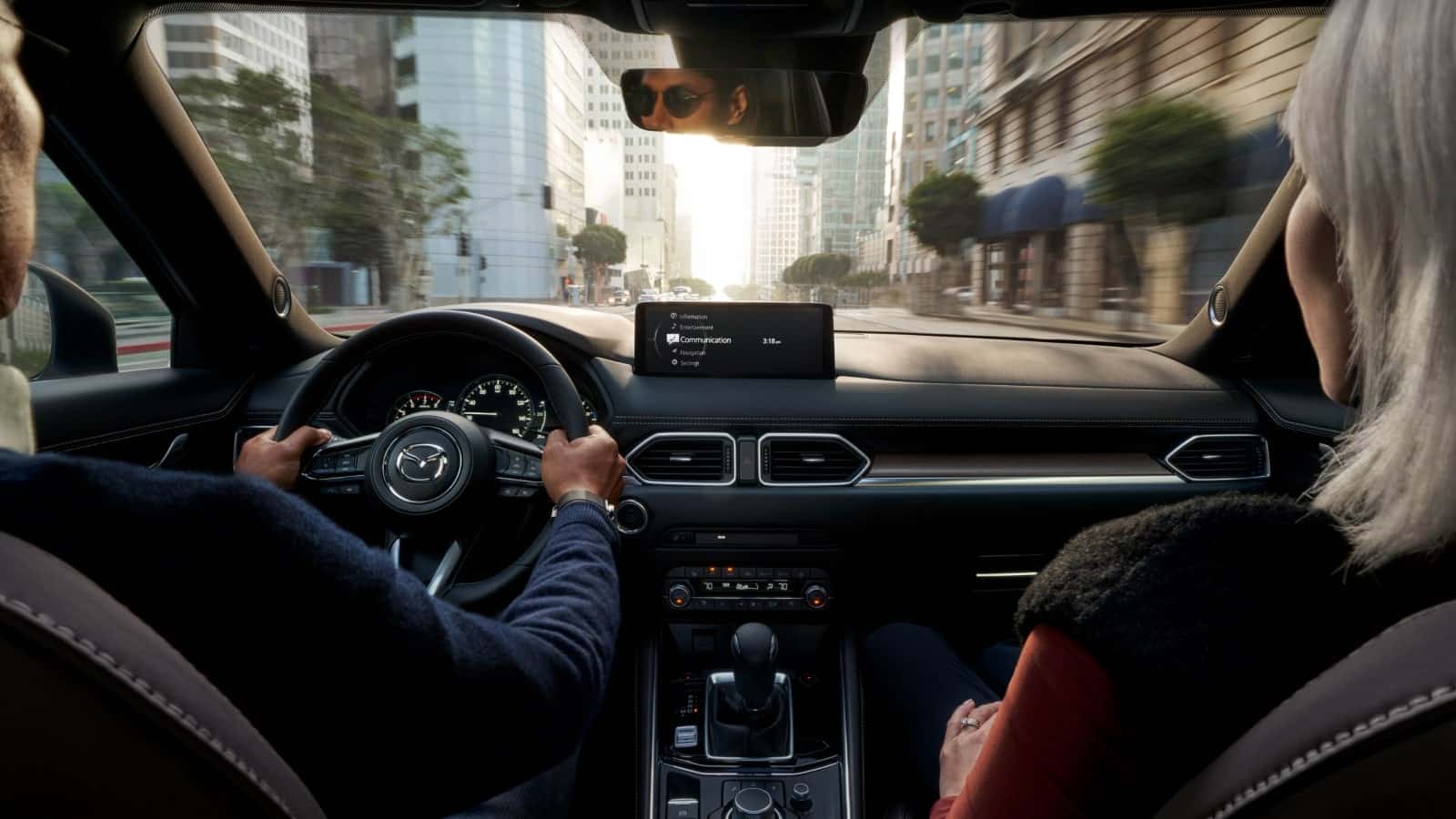 A couple is driving a 2025 Mazda CX-5 with hands on the wheel and showing instrument panel and gauges along with interior features while looking out of windshield.