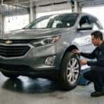 Technician kneeling beside the front tire of a gray Chevrolet SUV in a service bay