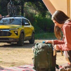 2025 Chevrolet Trailblazer in yellow parked at a campsite, with a man securing a bicycle on the roof rack and a woman preparing gear next to a tent.
