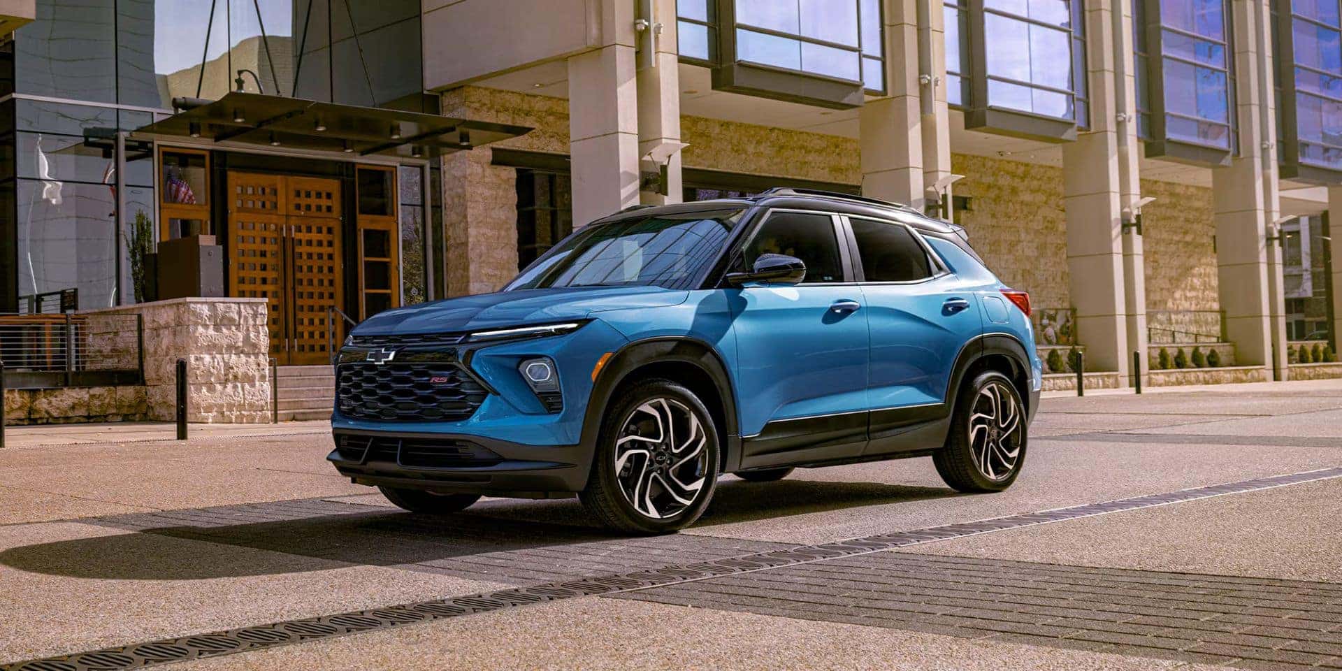 Front and side view of the 2025 Chevrolet Trailblazer in a striking blue finish, parked in front of a modern building with large windows and stone accents, highlighting its sporty design and alloy wheels.