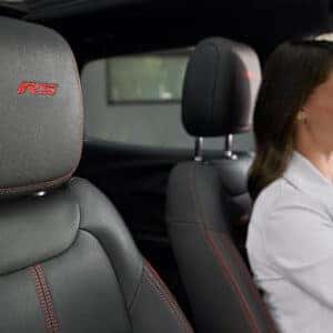 Close-up of the RS-embroidered front seat headrest in the 2025 Chevrolet Trailblazer, featuring premium black leather with red stitching, with a smiling driver in the background.