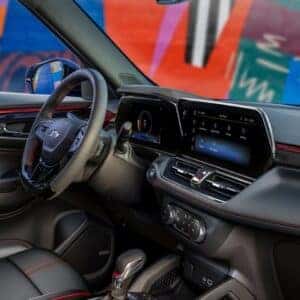 Driver's side interior view of the 2025 Chevrolet Trailblazer showcasing a stylish cabin with red accent trim, digital driver display, and infotainment system, complemented by a vibrant, artistic mural visible through the window.