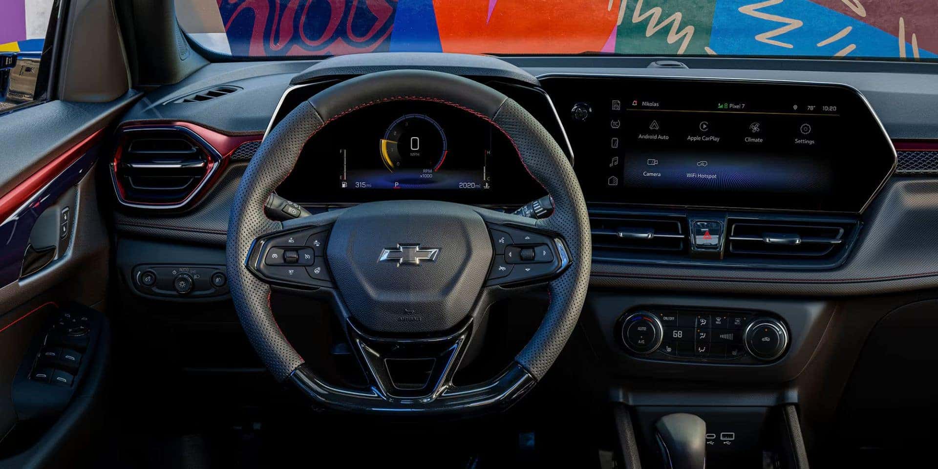 Close-up view of the 2025 Chevrolet Trailblazer's steering wheel with red stitching, digital driver display, and center console featuring a touchscreen infotainment system, set against a vibrant, artistic background visible through the windshield.