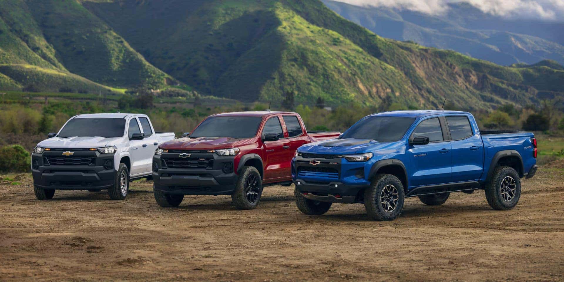 Three Chevrolet Colorado trucks in white, red, and blue colors parked side by side in a scenic valley with lush green mountains.