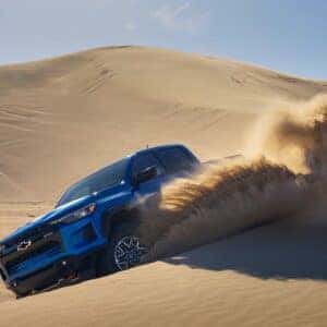 A blue Chevrolet Colorado driving through sand dunes, creating a cloud of dust.