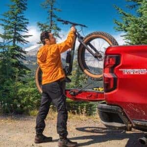 A man loading a mountain bike into the bed of a red Chevrolet Colorado Trail Boss in a forest setting.