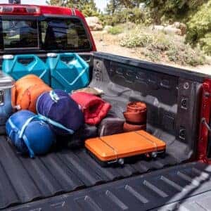 The bed of a red Chevrolet Colorado loaded with camping gear, including sleeping bags and water jugs, in a forested area.