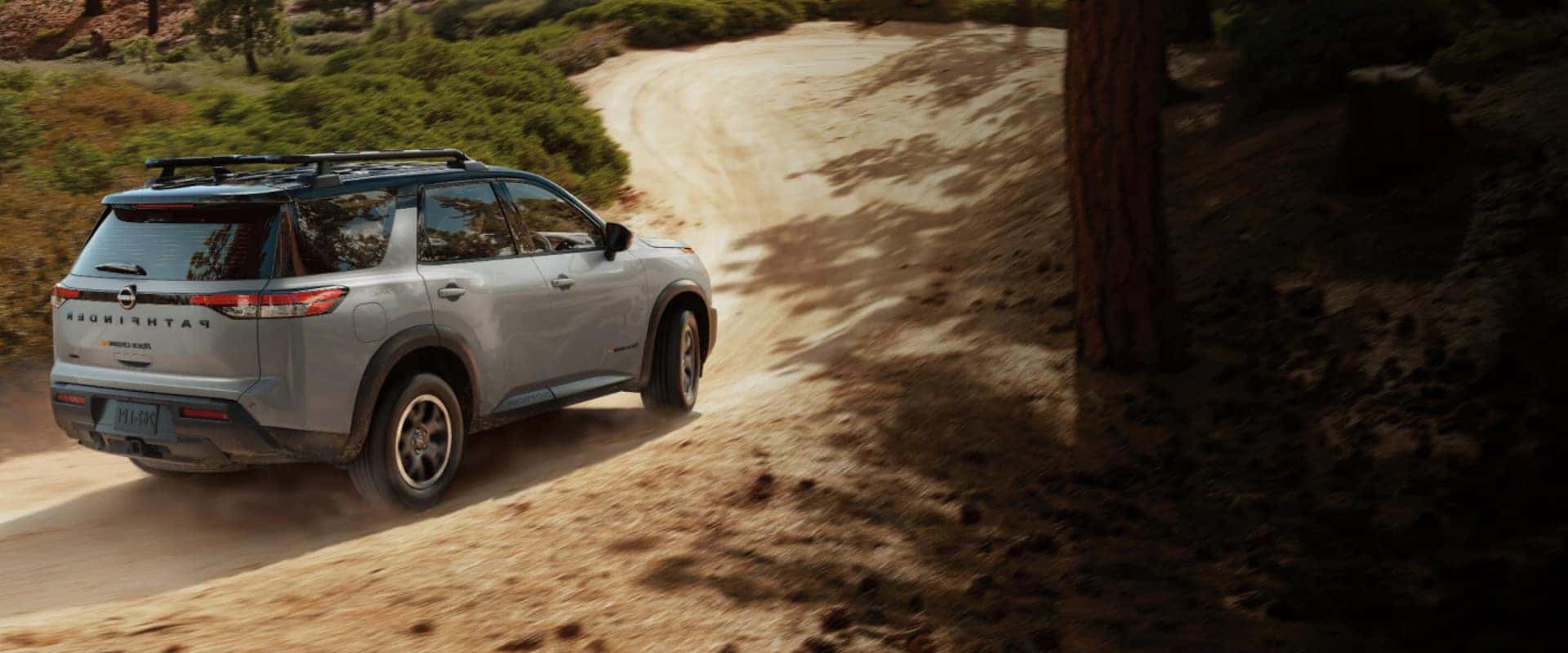 Exterior rear side hero image of a light gray 2025 Nissan Pathfinder Rock Creek driving up a dirt hill in a park landscape