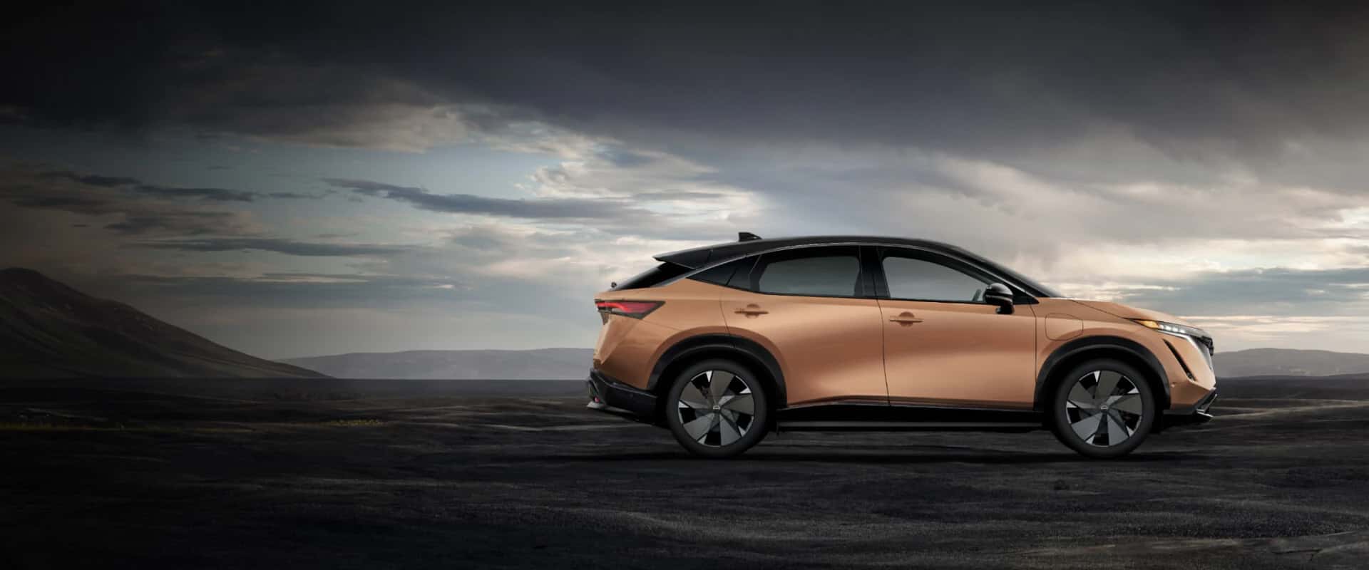 Exterior side hero image of a copper 2025 Nissan ARIYA parked in a dirt mountain landscape on a cloudy day