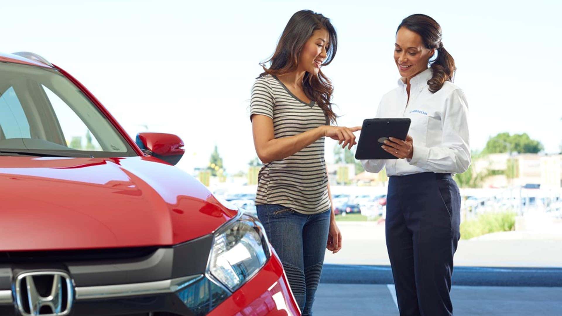 customer talking to woman at dealership with clipboard next to red vehicle