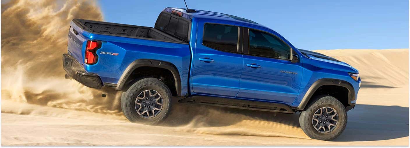 > Rear three-quarter view of a blue 2026 Chevrolet Colorado pickup truck kicking up a large cloud of sand while driving across sand dunes under a clear blue sky.