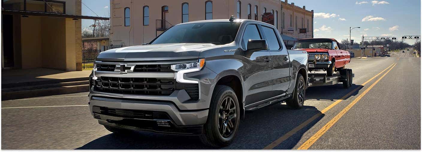 > Front three-quarter view of a gray 2026 Chevrolet Silverado 1500 pickup truck towing a flatbed trailer carrying a red classic car down a town street with historic buildings in the background.