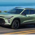 > Front three-quarter view of a light green 2026 Chevrolet Trax driving along a scenic coastal highway with a guardrail and the blue ocean in the background.