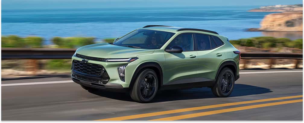 > Front three-quarter view of a light green 2026 Chevrolet Trax driving along a scenic coastal highway with a guardrail and the blue ocean in the background.