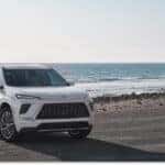 A white 2026 Buick Enclave SUV is parked on an asphalt turnout, viewed from a front three-quarter angle with a scenic background of ocean waves and coastal cliffs.