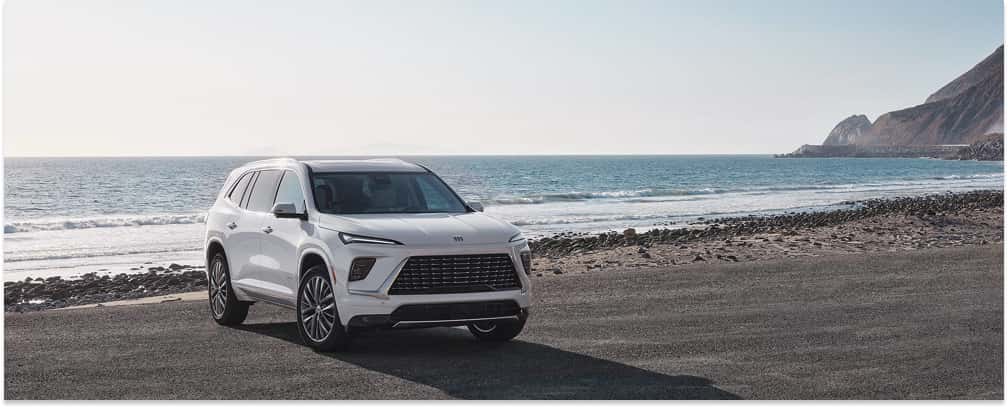 A white 2026 Buick Enclave SUV is parked on an asphalt turnout, viewed from a front three-quarter angle with a scenic background of ocean waves and coastal cliffs.