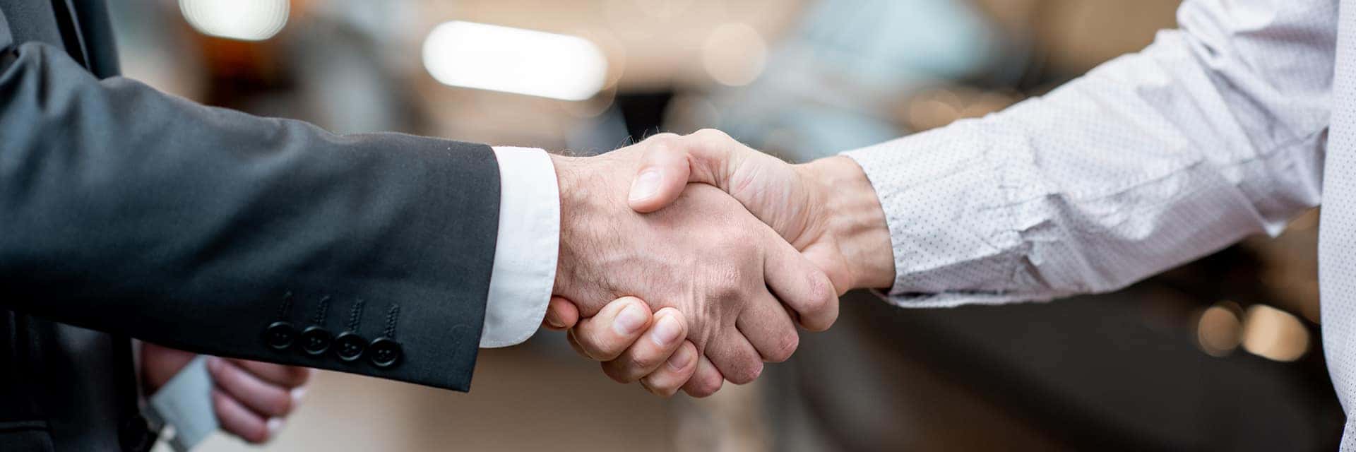 two people shaking hands in front of a vehicle at a dealership