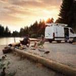 A White Sprinter Van being used at a campsite near a lake and two campers in folding chairs with geese nearby